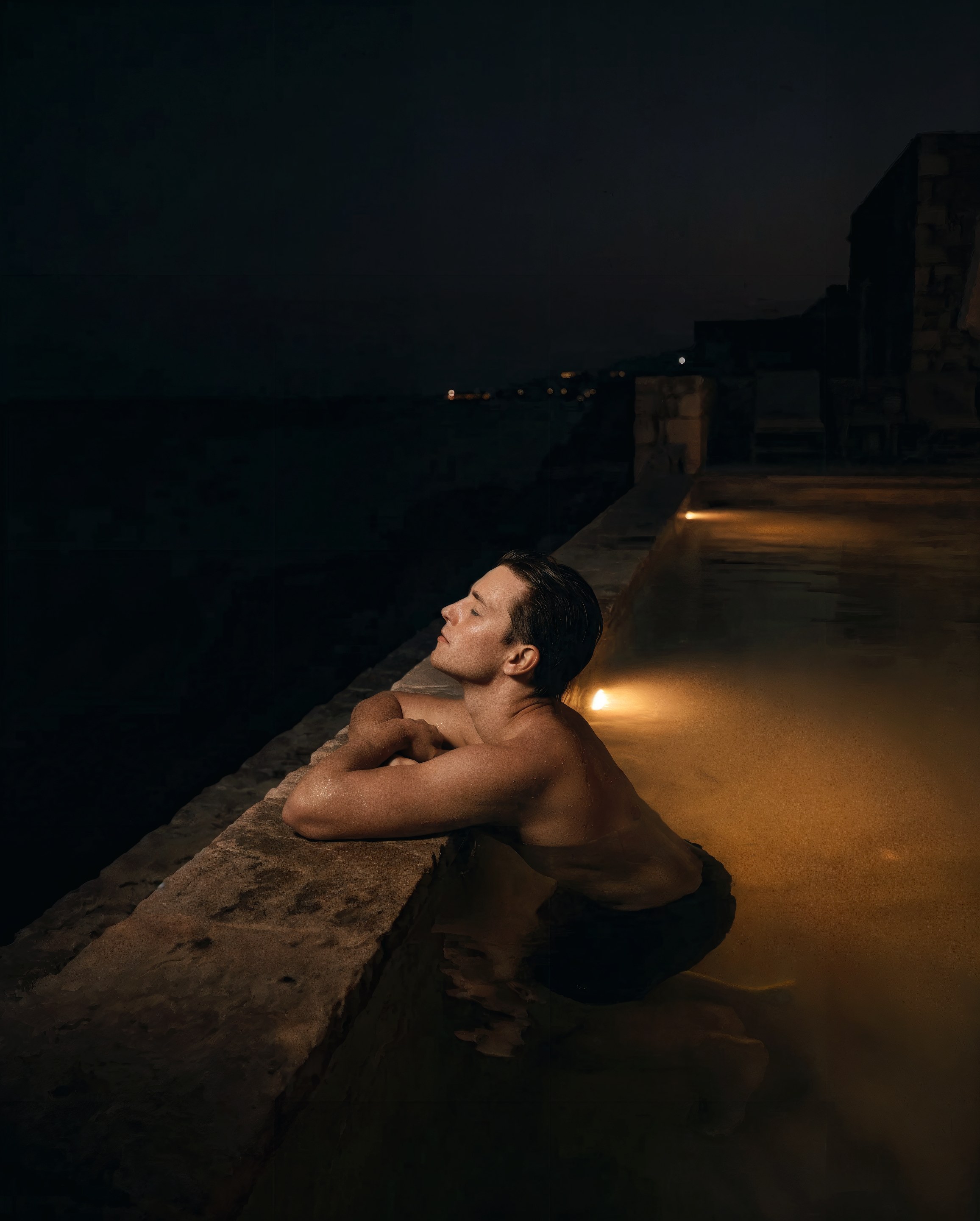 A man resting his arms on the edge of a dark infinity pool at night, lit from below by a single warm pool light, distant coastal lights beyond
